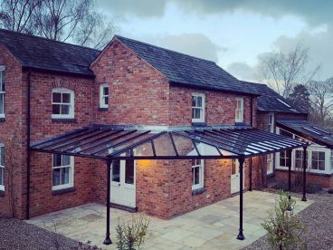 <p>We designed this unusual triangular shaped corner to follow the line of the existing patio on this stunning home in Leicestershire. The verandah provides around 30m2 of covered space, using our simple trumpet columns, with a glass roof and guttering included. Finished in black. </p>
